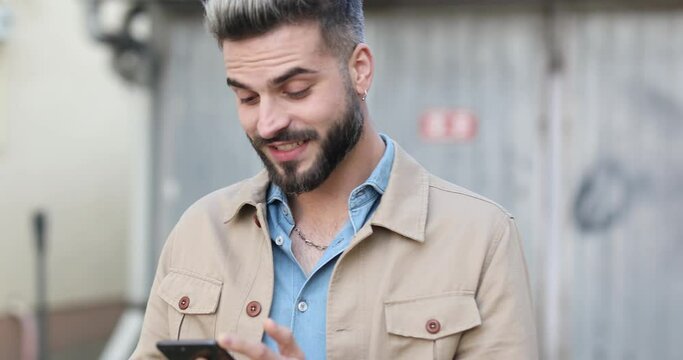 Happy Handsome Man In Denim Shirt Reading Emails And Answering Phone, Making Thumbs Up Gesture And Smiling In Front Of Gray Metal Garage Outside