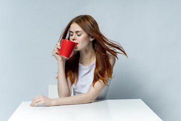 attractive red-haired woman drinking from a red mug while sitting at the table