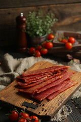 Delicious smoked sausage on a dark wooden board with tomatoes and spices. Delicious handmade snack made from natural ingredients. Pieces of dry pork, beef, chicken snacks. Meat products. Close-up.