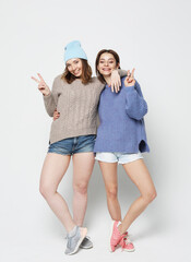 A full-length portrait of two smiling girls dressed in hipster style showing v-sign.
