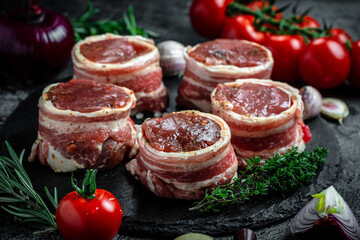 Raw pork medallion steaks wrapped in bacon served on a slate board on dark concrete background with spices salt and pepper, top view