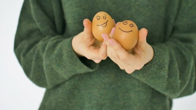 Young female in green sweater is showing two hand drawn eggs with anthropomorphic smiley face, easter preparation, holiday mood concepts