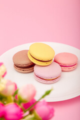 Colorful macaroons on white plate, pink background, flowers. Mothers, women day, Valentines day, birthday, spring, Easter. Copy space, vertical