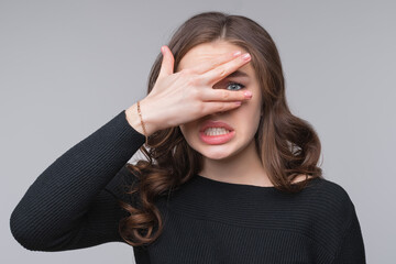 Close up worried, scared young woman looking between fingers, isolated over gray background
