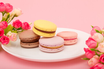 Plate of macaroons on pink background with flowers. Sweet pastry, baked products, sweets, dessert. Unhealthy diet, sugar dependence.