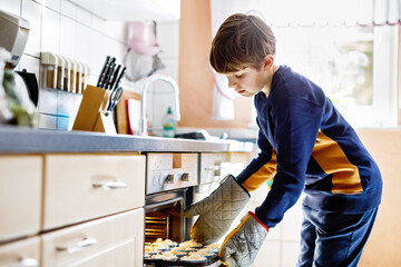 Happy blond school kid boy baking blueberry vanilla muffins in domestic kitchen, indoors. Funny...