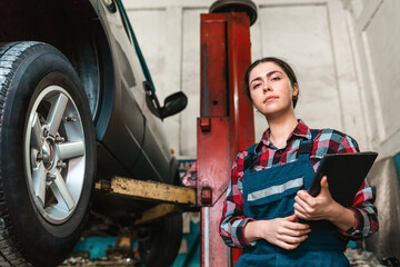 Portrait of a young female mechanic in uniform posing with a tablet in her hands. In the background is an auto repair shop. Bottom view