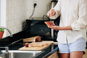 Cropped image of young woman making sweet sandwich and spreading berry jam on bread