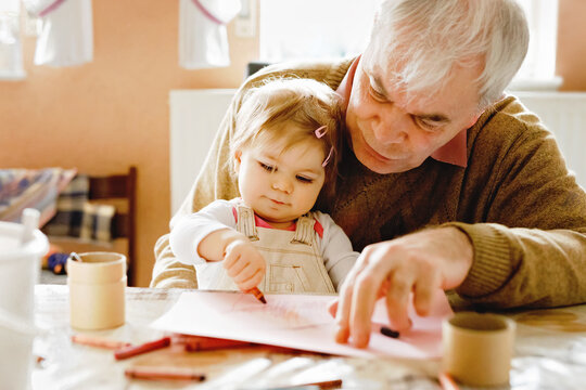 Cute little baby toddler girl and handsome senior grandfather painting with colorful pencils at home. Grandchild and man having fun together. Family and generation in love