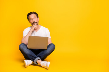Full size photo of serious minded man sit floor hold laptop look empty space isolated on yellow color background