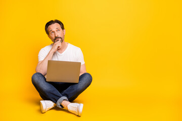 Full size photo of serious minded man sit floor hold laptop look empty space isolated on yellow...