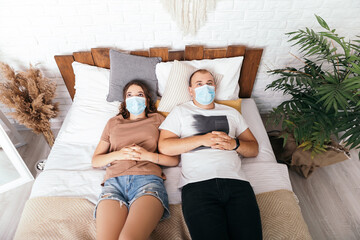 Obraz premium Young lovely couple lying on bed boring mood wear face disposable blue mask, apartment interior. Contemporary studio flat. Husband and wife lovers, coronavirus concept copy space 
