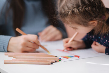 Adirable little girl with her beautiful young mom draws in the album using color pensins. Cute girl paints coloring page at home during quararntine. Happy kid sits at the table by the window. Closeup