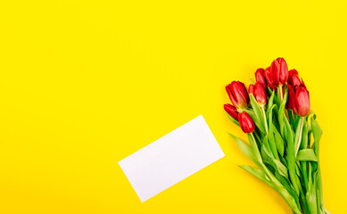 White gift certificate envelope or business card on yellow background with bouquet of red tulips and copy space, empty text place. Holiday greeting card. Happy Valentine Day, Woman Day, Mother Day