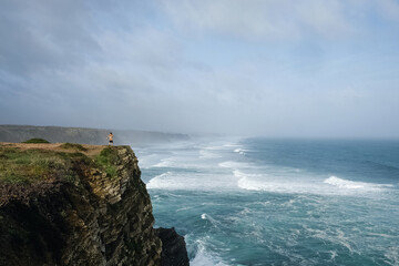Small unrecognizable figure of woman standing on a cliff at ocean on windy day