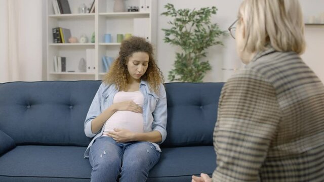 Blond psychoanalyst talking to sad expecting female, sitting on the sofa, advice
