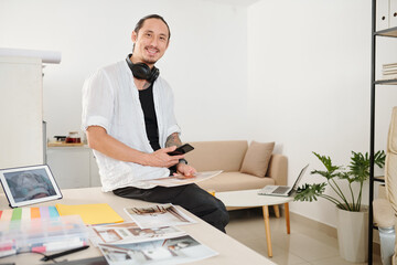 Fototapeta na wymiar Portrait of happy interior designer sitting on table edge and checking printed photos of house or apartment