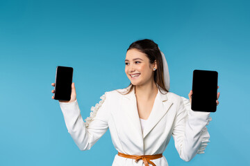 Naklejka premium Happy girl in a white jacket with a smile shows two smartphones at the camera. Black blank screens. Blue background. Advertising space.