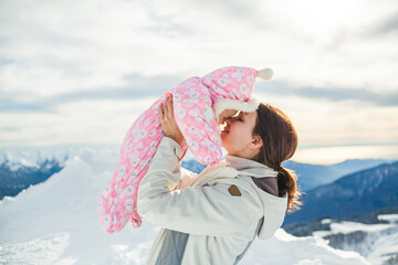 Fototapeta na wymiar Young woman kisses her little daughter at sunny snowy slope. Young mother embrace her baby on the mountain top. Mom and cute baby having good time in the winter nature. Mothers Day. Family Concept