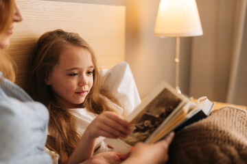 Close-up of mother and happy daughter reading together children book before going to sleep while lying in bed in nursery bedroom, near lamp. Concept of family leisure activity at home.