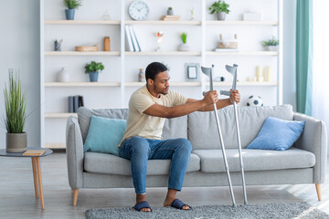 Young black guy having difficulty standing up from sofa, leaning on crutches at home