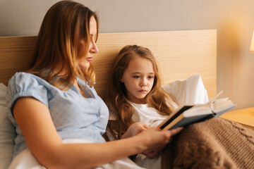 Side view of young happy mother and adorable cheerful daughter reading children book together lying...