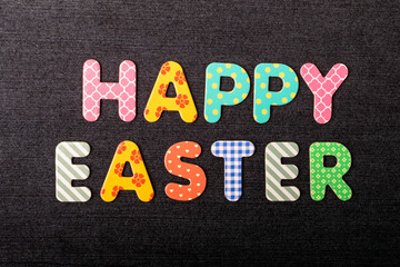 Card with Happy Easter words made from mixed vivid colored wooden letters on a textured dark black textile material that can be used as a message.