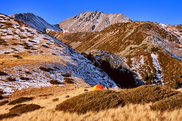 Tent set up on the ridge of mountain in the highlands on the background of blue sky, bright orange tent fluttering in the wind; travel and tourism concept