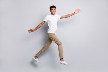 Fototapeta na wymiar Photo of cheerful guy jump raise hands look camera toothy smile wear white t-shirt pants sneakers isolated grey background