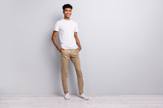 Full size photo of brunet optimistic curly guy stand wear white t-shirt trousers sneakers isolated on grey color background