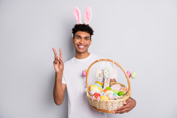 Photo of guy hold easter basket show v-sign wear bunny ears headband white t-shirt isolated grey...