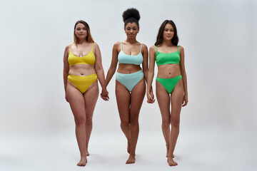 Full length shot of three confident diverse women in colorful underwear holding hands, looking at camera, standing together isolated over light background - obrazy, fototapety, plakaty