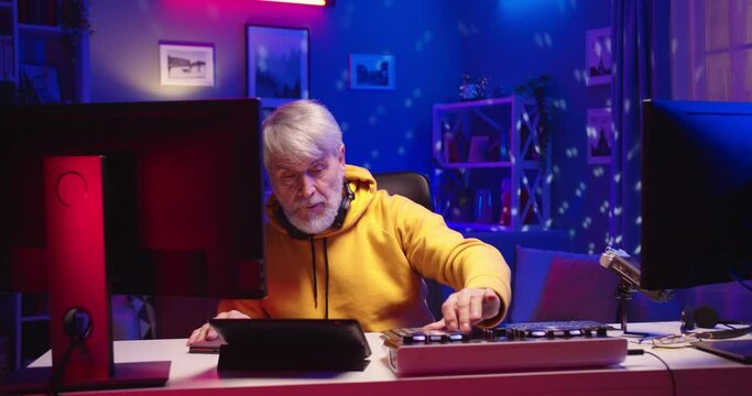 Caucasian concentrated old man professional DJ working in home record studio use surface control desk equalizer mixer, colorful neon lights, senior musician streaming live on computer Portrait concept