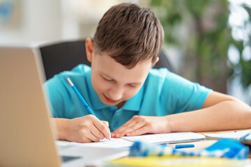 inquisitive boy uses laptop to study school subjects during his online lesson at home. Writing down topic lesson in notebook. Selective focus on the child's hand. E-Education Distance Home Education.