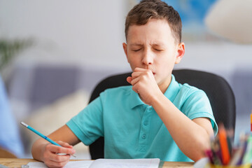 small child, schoolboy of 8 years old, is studying at a desk with laptop, at home. School distance education at home during quarantine concept. Coughing, sneezing, covering your mouth with your hand.