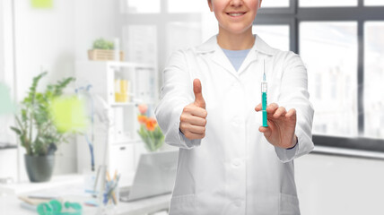medicine, vaccination and healthcare concept - close up of happy smiling female doctor or nurse with syringe showing thumbs up over medical office at hospital background