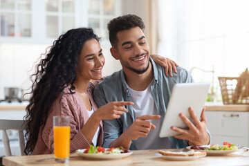 Fototapeta premium Cheerful Middle Eastern Spouses Using Digital Tablet While Having Breakfast In Kitchen