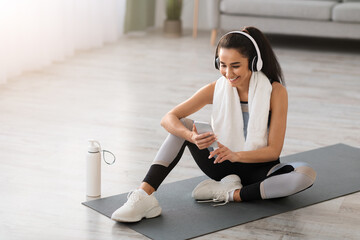 Happy young woman resting on fitness mat, listening to music