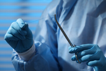 Professional surgeon holding forceps with suture thread on blurred background, closeup. Medical...