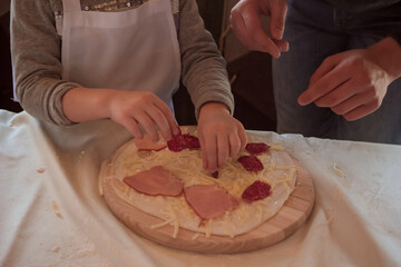 Cooking class, culinary. Kids hand puts ingredients on a row pizza on a round wooden cutting board sprinkled with flour on the white table. Children in uniform preparing food, meal in the kitchen