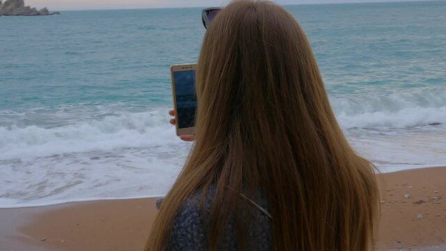 Young red hair woman takes a shot of the blue Adriatic sea staying on the beach with smartphone. Wild nature in Montenegro, southern europe