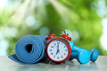 Alarm clock, yoga mat and dumbbells on marble table outdoors. Morning exercise