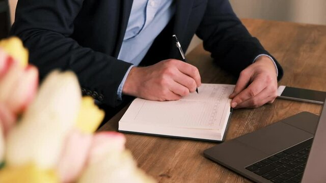 Side view of businessman in suit filling out planner or notepad next to an open laptop against of fresh bouquet of tulips. A manager or white collar worker is practicing time management. Slow motion