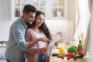 Cheerful Arab Couple Using Digital Tablet While Cooking Healthy Lunch In Kitchen