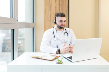 Man professional doctor in white medical coat and headset making conference call on laptop...