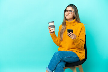 Young caucasian woman sitting on a chair isolated on blue background holding coffee to take away and a mobile while thinking something