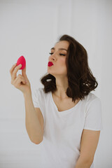 Portrait of beautiful young woman is having fun with pink egg sponge. Isolated over white background. Copy space. Skin care and beauty concept.