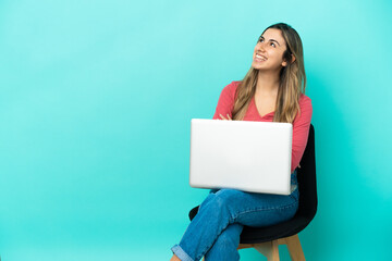 Young caucasian woman sitting on a chair with her pc isolated on blue background looking up while smiling