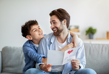 Happy father's day. Boy embracing dad and congratulating him with handmade postcard, smiling to each other