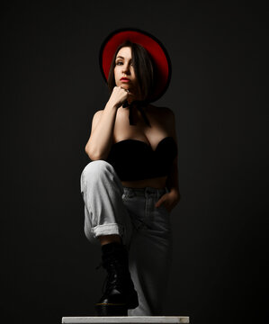 Sensual sexy brunette woman in massive boots, black top bra, jeans and hat stands stepping on chair holding fist at chin over black background. Fashion, stylish casual look for woman concept